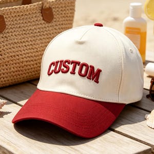 Peut inclure: Une casquette de baseball avec une calotte crème et une visière rouge. Le mot CUSTOM est brodé en lettres capitales rouges sur le panneau avant. La casquette présente une structure rigide avec un bouton sur le dessus et des coutures visibles.
