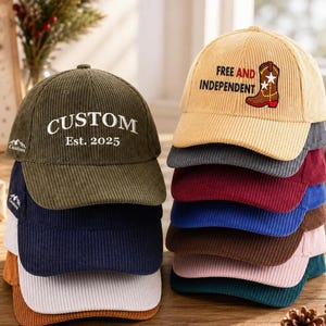 Puede incluir: Una pila de gorras de béisbol de pana en varios colores. La gorra verde oliva tiene el texto CUSTOM Est. 2025. La gorra color canela presenta un gráfico de bota vaquera con el texto FREE AND INDEPENDENT. Cada gorra tiene una visera curva.