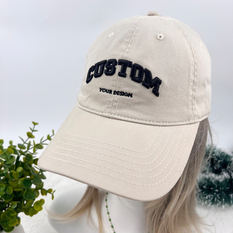 May include: A beige baseball cap with a curved brim. The cap features the word "CUSTOM" in large, black, raised letters above the text "YOUR DESIGN". The cap is made of a soft fabric and has a classic six-panel design.