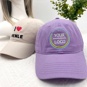 May include: Two baseball caps. One is tan with the text "I ❤️ HENLE" embroidered in black and red. The other is purple with a circular logo that says "YOUR LOGO" in white, with blue and yellow accents.