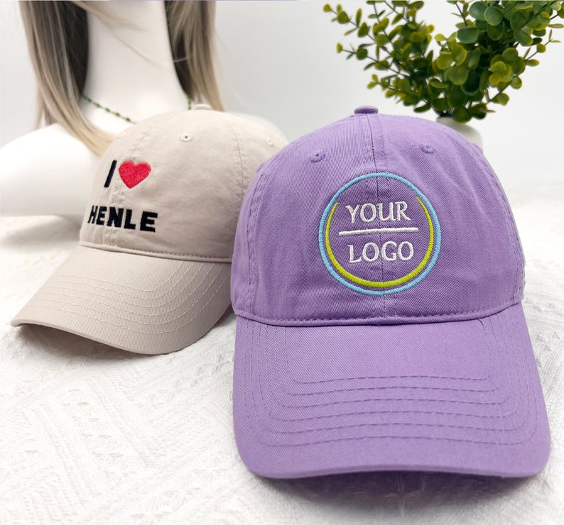 May include: Two baseball caps. One is tan with the text "I ❤️ HENLE" embroidered in black and red. The other is purple with a circular logo that says "YOUR LOGO" in white, with blue and yellow accents.