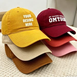 May include: A stack of baseball caps in various colors, including mustard yellow, burgundy, and pink. The yellow cap has the text "YOUR DESIGN HERE" in white. The burgundy cap has the text "MY FAVORITE CONTO OMTSO" in white.