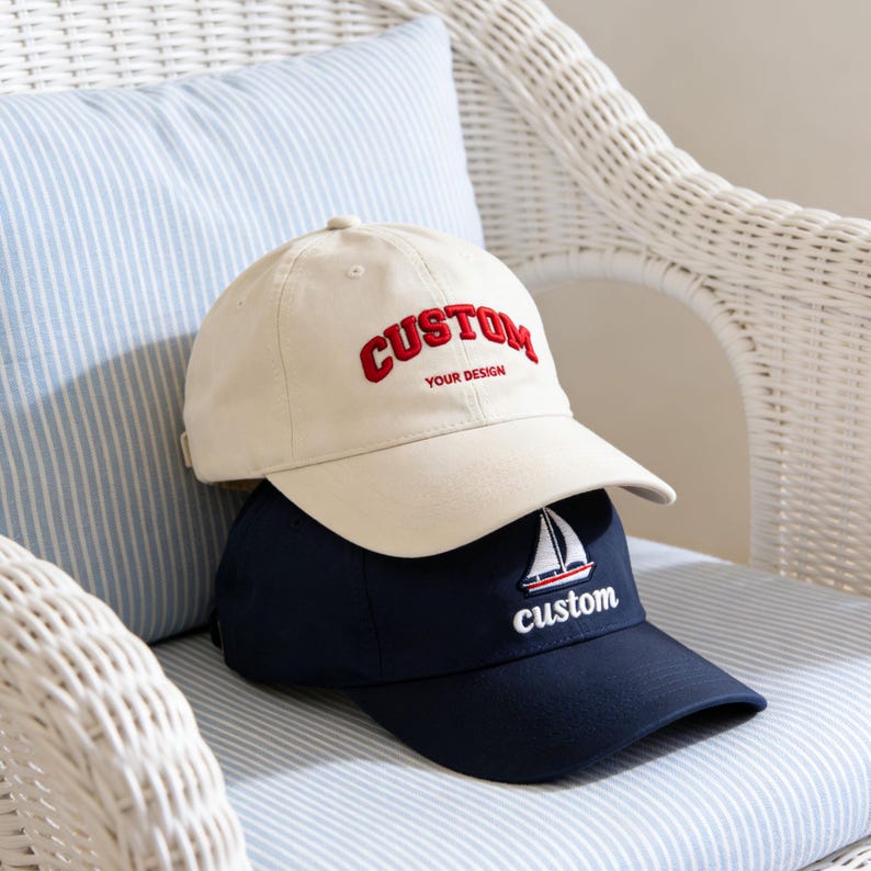 May include: Two baseball caps stacked on a chair. The top cap is beige with the word "CUSTOM" in red script and the text "YOUR DESIGN" below. The bottom cap is navy blue with a sailboat graphic and the word "custom".