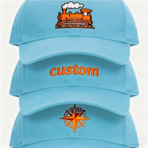 May include: A stack of light blue baseball caps. The top cap features an orange train embroidery. The middle cap has the word "custom" embroidered in orange. The bottom cap has a compass rose design.