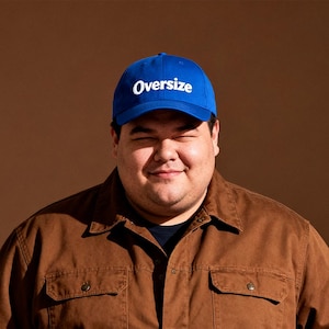 May include: A blue baseball cap with the word "Oversize" in white lettering. The cap is worn by a person wearing a brown button-up shirt.