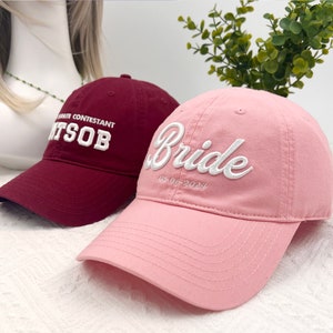 May include: Two baseball caps. One is burgundy with white embroidered text reading "FAVORITE CONTESTANT ITSOB". The other is pink with "Bride" embroidered in white, with the date "05-06-2024" below.