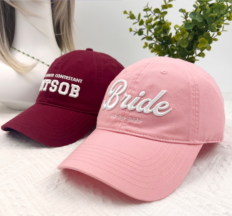 May include: Two baseball caps. One is burgundy with white embroidered text reading "FAVORITE CONTESTANT ITSOB". The other is pink with "Bride" embroidered in white, with the date "05-06-2024" below.
