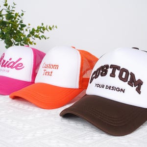 May include: Three trucker hats with mesh backs. The first hat is pink with the word "Bride" embroidered in white. The second hat is orange with "Custom Text" embroidered in white. The third hat is brown with "CUSTOM YOUR DESIGN" embroidered in white.