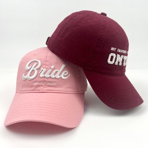 以下が含まれることがあります： 2つのベースボールキャップ。1つはピンクで「Bride」と「05-06-2024」が白で刺繍されており、もう1つはマルーンで「My Favorite OM」が白で刺繍されている。