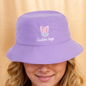 May include: A lilac bucket hat featuring a floral design and the words "Custom logo". The hat is made of a soft fabric and has a wide brim. The hat is being worn by a person.
