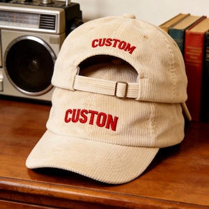 Op de afbeelding: Twee beige corduroy baseballpetten, op elkaar gestapeld. Het woord "CUSTOM" is rood geborduurd op de bovenste pet en "CUSTON" op de onderste pet. De petten hebben een gebogen klep en een verstelbare band.