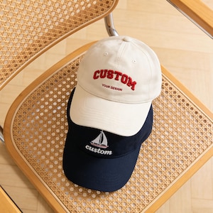 May include: Two baseball caps, one beige and one navy blue, are stacked on a wooden chair. The beige cap has "CUSTOM" embroidered in red, with "YOUR DESIGN" below. The navy cap features a sailboat graphic and the word "custom".