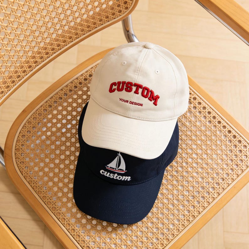 May include: Two baseball caps, one beige and one navy blue, are stacked on a wooden chair. The beige cap has "CUSTOM" embroidered in red, with "YOUR DESIGN" below. The navy cap features a sailboat graphic and the word "custom".