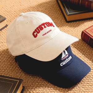 Könnte beinhalten: Zwei Baseballkappen, eine cremefarbene und eine marineblaue, sind übereinander gestapelt. Die cremefarbene Kappe hat "CUSTOM" in Rot mit "YOUR DESIGN" darunter gestickt. Die marineblaue Kappe zeigt ein Segelboot und das Wort "custom".