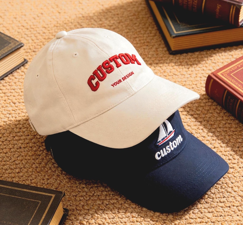 May include: Two baseball caps, one cream-colored and one navy blue, are stacked. The cream cap has "CUSTOM" embroidered in red with "YOUR DESIGN" below. The navy cap features a sailboat and the word "custom".