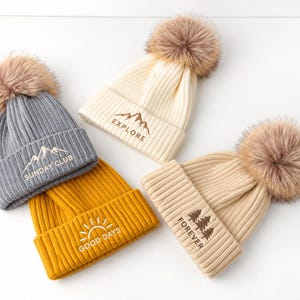 Op de afbeelding: Vier geribbelde gebreide mutsen met imitatiebont pompons in grijs, crème, mosterdgeel en beige. Elke muts heeft geborduurde ontwerpen: een berg met 'SUNDAY CLUB', een berg met 'EXPLORE', een zon met 'GOOD DAYS' en bomen met de tekst 'FOREVER'.