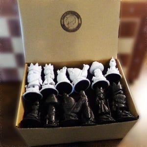 May include: A chess set with black and white pieces, displayed in a cardboard box. The pieces are stylized, with white figures on black bases. The box has a circular emblem on the top.