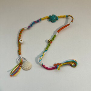 May include: A colorful friendship bracelet with a turquoise turtle charm, a white seashell, and gold beads. The bracelet has a ring attached to it and is tied with tassels in various colors.