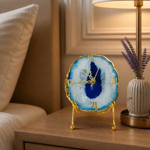 May include: A decorative agate clock with a blue and white center and gold-colored hands and frame. The clock is displayed on a gold stand. Roman numerals mark the hours. The clock is on a wooden nightstand next to a lamp.