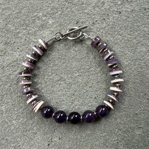 May include: A bracelet featuring a toggle clasp, alternating between flat purple and white striped disc beads, small metallic silver spacer beads, and five smooth, round, deep purple gemstone beads arranged at the bottom of the circular design.