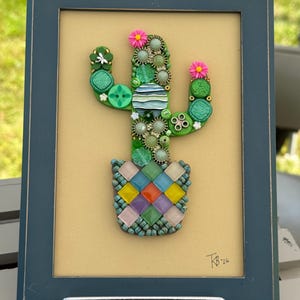 May include: A framed mosaic art piece featuring a cactus made of various green buttons, beads, and two pink flowers. The cactus sits in a pot constructed from small, multi-coloured square tiles. The signature KB '26 is visible in the bottom right corner.