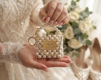 Puede incluir: Un bolso en miniatura hecho de perlas artificiales blancas con una placa de metal dorado grabada con el texto Büşra & Resul 07.09.2026. El bolso cuenta con un asa de perlas, un llavero de metal dorado y una cadena decorativa dorada.