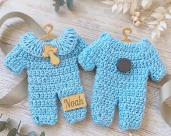 Puede incluir: Dos peleles de bebé tejidos a ganchillo en color azul claro colgados de pequeñas perchas doradas. El pelele de la izquierda tiene un dije de chupete dorado y una etiqueta rectangular dorada con el texto Noah. El de la derecha tiene un botón circular oscuro.