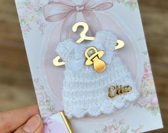 Puede incluir: Un adorno de vestido blanco tejido a ganchillo en una percha dorada, con un dije de chupete dorado y el nombre Eliz en letra dorada. El adorno está sujeto a una tarjeta floral con un lazo, junto a una pequeña barra de chocolate Baby Eliz.