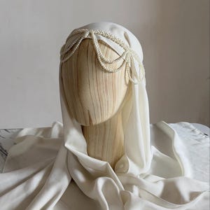 May include: A white head covering draped over a wooden mannequin head, featuring a decorative headpiece made of multiple strands of small white pearls arranged in a scalloped pattern across the forehead and sides of the head.