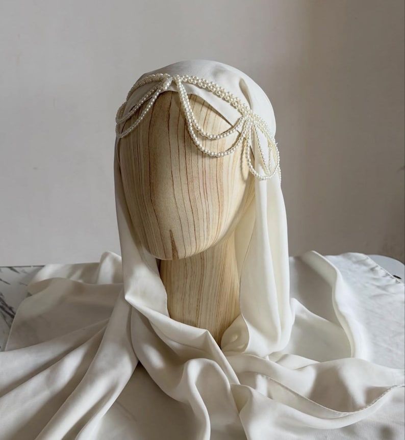 May include: A white head covering draped over a wooden mannequin head, featuring a decorative headpiece made of multiple strands of small white pearls arranged in a scalloped pattern across the forehead and sides of the head.