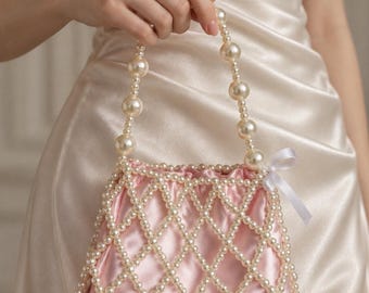 Puede incluir: Un bolso de mano de satén rosa claro con un revestimiento exterior de red de perlas artificiales blancas. El bolso incluye un asa superior hecha de grandes perlas artificiales blancas y un pequeño lazo de cinta blanca en el lateral.