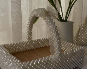 Puede incluir: Una cesta rectangular con base de madera clara y paredes formadas por filas de perlas blancas. El asa está adornada con un lazo de cinta de organza blanca transparente. Una pequeña etiqueta rectangular en el lateral muestra el texto Lila Beads.