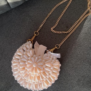 May include: A round, peach-coloured beaded handbag with a gold chain strap. The bag features a floral pattern and a small ribbon. The chain is attached to the bag with gold-coloured clasps.