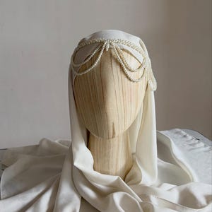 May include: A cream-colored head covering draped over a wooden mannequin head, featuring a decorative headpiece made of multiple strands of small white pearls arranged in a layered, scalloped pattern across the forehead and sides.