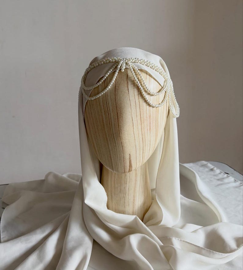 May include: A cream-colored head covering draped over a wooden mannequin head, featuring a decorative headpiece made of multiple strands of small white pearls arranged in a layered, scalloped pattern across the forehead and sides.