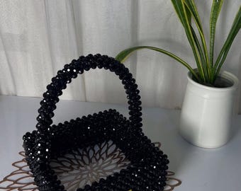 Puede incluir: Una cesta de forma cuadrada construida con pequeñas cuentas negras facetadas. La cesta cuenta con un asa arqueada única hecha de las mismas cuentas negras e incluye una pequeña etiqueta metálica dorada en la parte frontal.