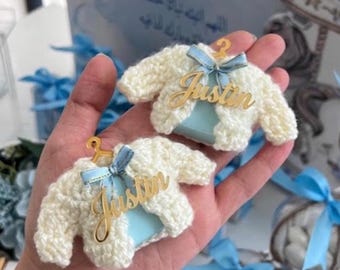 Puede incluir: Dos suéteres en miniatura de ganchillo color crema, cada uno colgado de una pequeña percha dorada. Una cinta azul claro está atada en el cuello de cada suéter, y una placa dorada con el nombre Justin en cursiva está fijada en la parte delantera.