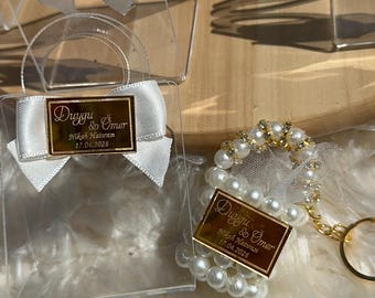 Puede incluir: Bolsas de regalo transparentes de acrílico con lazos de satén blanco y etiquetas doradas. Una bolsa tiene un asa y otra tiene un llavero de perlas y pedrería. Las etiquetas dicen "Duygu & Ömer Nikah Hatırası 17.04.2026".
