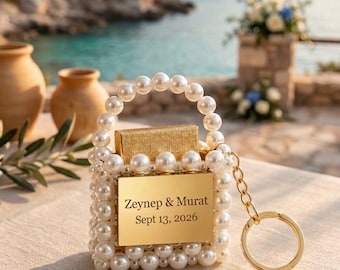 Puede incluir: Un mini bolso cuadrado cubierto de perlas artificiales blancas con un asa de perlas y una placa de metal dorado en la parte frontal. La placa tiene grabado el texto Zeynep & Murat Sept 13, 2026. Incluye una cadena de metal dorado con un llavero.