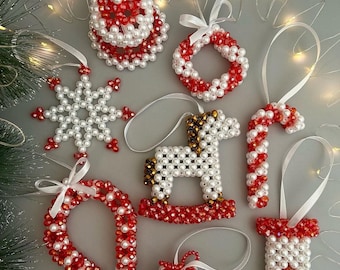 Puede incluir: Una colección de adornos navideños hechos a mano con cuentas blancas tipo perla y cuentas de cristal rojo facetado. El conjunto incluye un copo de nieve, un caballito balancín, un bastón de caramelo, un calcetín, una corona, una campana y un regalo.