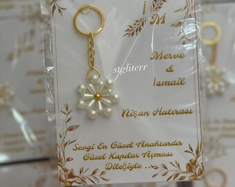 Puede incluir: Un llavero dorado con un colgante blanco en forma de flor hecho de perlas y cuentas. El llavero está unido a una tarjeta blanca con detalles florales dorados y el texto "Merve & İsmail" y "Nişan Hatırası".