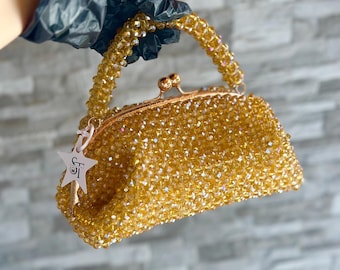 Puede incluir: Un bolso de mano dorado con cuentas, cierre de broche y asa de cuentas. El bolso tiene una etiqueta en forma de estrella con las iniciales "ST". El bolso lo sostiene una persona con un guante negro.