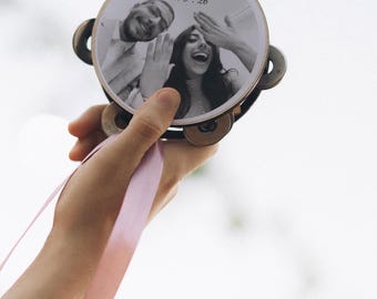 Puede incluir: Una pandereta redonda de madera con una fotografía en blanco y negro de dos personas y el texto Yassin & Jana 26.6.26 impreso en la superficie. El instrumento tiene sonajas de metal alrededor del marco y una cinta rosa claro a un lado.