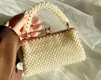 Puede incluir: Un bolso de mano rectangular completamente cubierto de pequeñas perlas artificiales de color crema. El bolso cuenta con un asa superior a juego hecha de perlas y un marco de metal dorado con un cierre de broche en la parte superior.