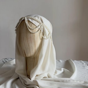 May include: A white head covering draped over a wooden mannequin head, featuring a decorative headpiece made of multiple strands of small white pearls arranged in a scalloped pattern across the forehead and sides of the head.