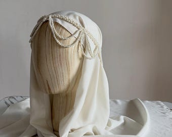 Puede incluir: Un cubrecabezas blanco drapeado sobre una cabeza de maniquí de madera, con un adorno compuesto por múltiples hilos de pequeñas perlas blancas dispuestas en un patrón festoneado sobre la frente y los lados de la cabeza.