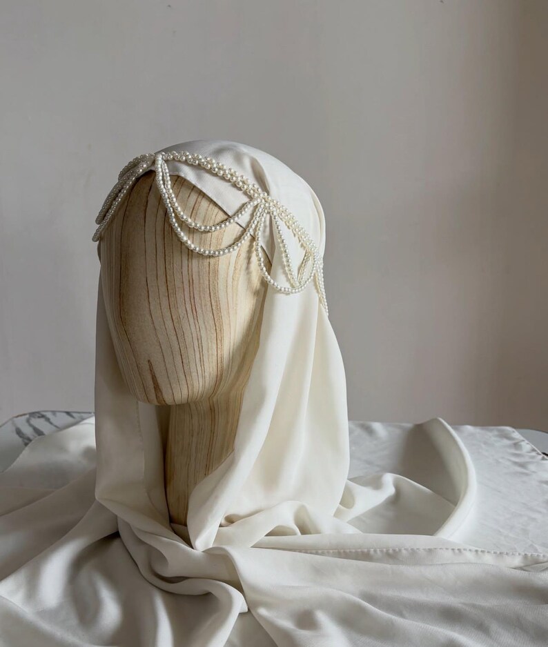 May include: A white head covering draped over a wooden mannequin head, featuring a decorative headpiece made of multiple strands of small white pearls arranged in a scalloped pattern across the forehead and sides of the head.