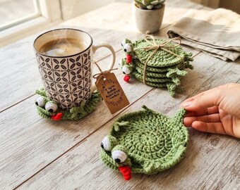 Puede incluir: Un juego de posavasos redondos de ganchillo verde con forma de rana, con ojos blancos y negros y lenguas rojas. Un posavasos está bajo una taza de cerámica estampada, mientras que otros cuatro están atados con un cordel de yute.