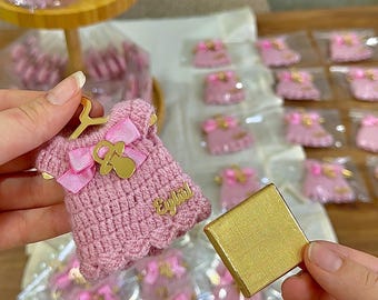Puede incluir: Un pequeño adorno de vestido tejido en rosa sobre una percha dorada en miniatura, con un lazo de satén rosa, un dije de chupete dorado y una placa dorada con el nombre Eylul, junto a una pieza de chocolate cuadrada envuelta en oro.