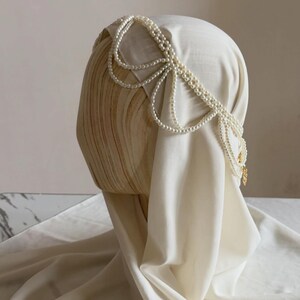 May include: A cream-colored head covering featuring an intricate arrangement of draped white pearl strands. The pearls are layered in multiple loops across the top and sides, accented with a small gold-toned metal charm at the end.
